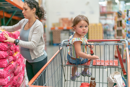 Adorable Eurasian toddler girl looking directly at the camera while seated in a shopping cart in a warehouse grocery store. In the background the child's pregnant mom is selecting an item to purchase.