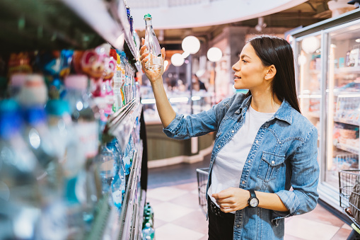 Happy young woman shopping in supermarket chooses water holding bottle standing near shelf