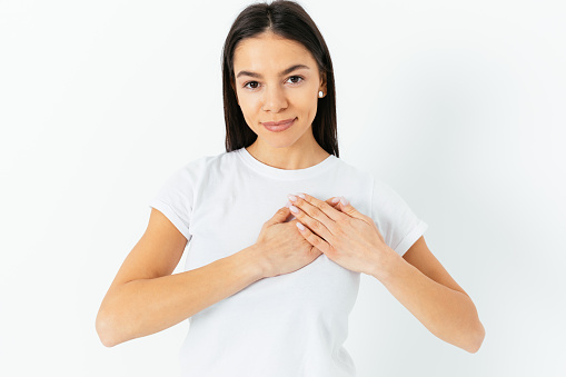 Friendly young woman with calm face and slight smile presses her hands to her chest feeling gratitude, looks with appreciation standing on white background