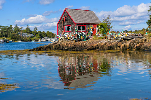 Bailey Island, Maine, USA - September 26, 2021: A small rustic fishing shack, surrounded by well-weathered but still colorful fishing floats, standing on rocky shore of Mackerel Cove of Bailey Island on a sunny Autumn morning. Note: Focus is on the shack.
