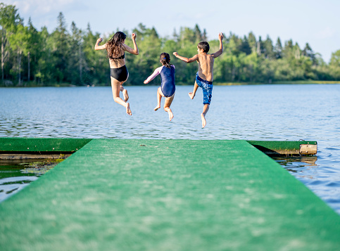 A small group of three children are seen in mid air jumping off the end of a dock on a sunny summer day. They are each striking a fun pose before they hit the waters surface to make a splash.