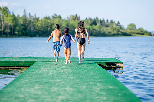 A small group of three children are seen running towards the end of a dock on a sunny summer day. They are each wearing swimsuits as they make their way to the end and prepare to jump off.