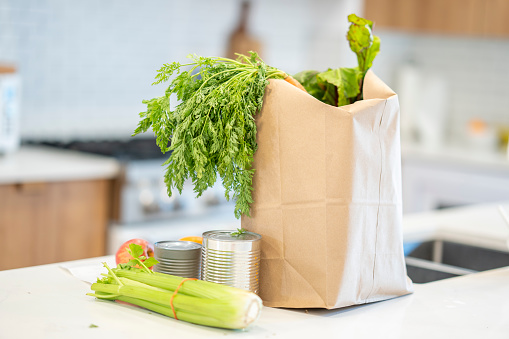 A paper bag of fresh produce sits on a kitchen counter after being purchased.  The leafy greens can be seen popping out of the tip of the bag and some vegetables are out on the counter beside the bag.