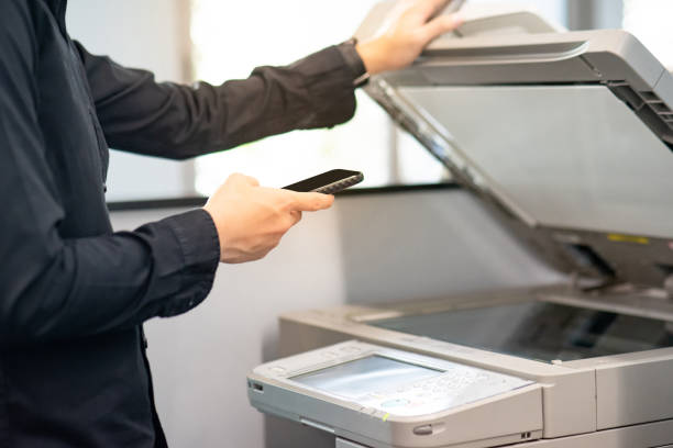 Using mobile app with photocopier Using mobile app with photocopier. Male hand holding smartphone while printing paperwork on photocopier. Electronic equipment and supply for business organization. computer printer office printout digital display stock pictures, royalty-free photos & images