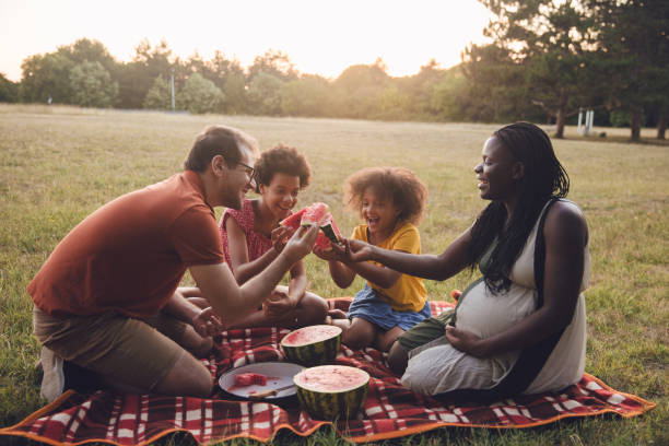 Multiracial family going for a picnic in a public park Multiracial family going for a picnic in a public park, In this photo, they enjoy eating a watermelon. His wife is in a late pregnancy period, close to delivering. black man eating watermelon stock pictures, royalty-free photos & images