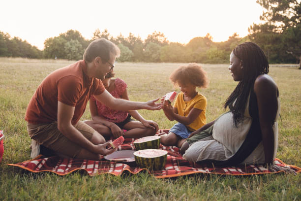 Multiracial family going for a picnic in a public park Multiracial family going for a picnic in a public park, In this photo, they enjoy eating and sharing a watermelon. His wife is in a late pregnancy period, close to delivering. black man eating watermelon stock pictures, royalty-free photos & images