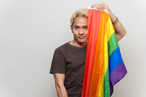 Studio portrait photo of young Asian man holding LGBTQ rainbow flag and looking at camera on white background. Young gay man emotion face expression portrait and LGBTQ people lifestyle concept.