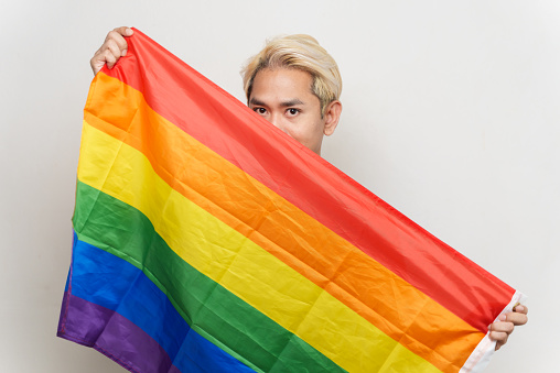 Studio portrait photo of young Asian man holding LGBTQ rainbow flag and looking at camera on white background. Young gay man emotion face expression portrait and LGBTQ people lifestyle concept.