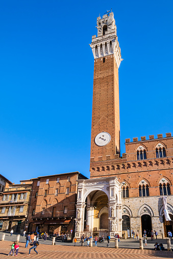Tourists strolling in the famous Piazza del Campo of Siena, where overlook several historic buildings, including the Torre del Mangia/Civic Tower, built in 14th century, and the Palazzo Pubblico, seat of the Town Hall.