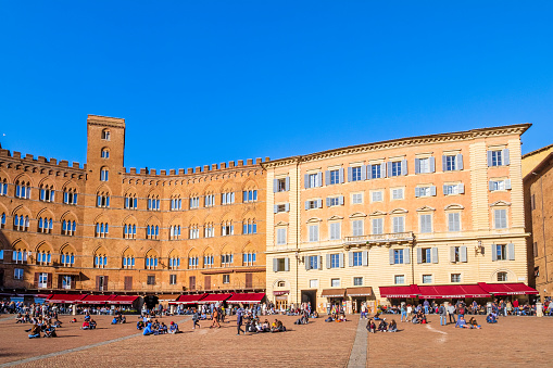 Tourists strolling in the famous Piazza del Campo of Siena, where overlook several historic buildings now hosting bars and restaurants.