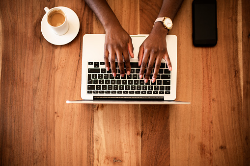 Overview of a female working on a laptop. She has coffee cup on one side and smart phone on another.