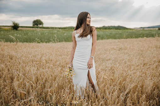 Beautiful young bride standing on a field in nature alone and holding a bouquet.