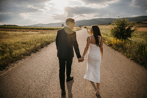 Groom and walking and holding hands bride on the middle of the road in nature.