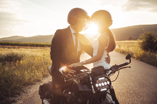 Groom sitting on his motorcycle and bride is standing next to him on the middle of the road in sunset.