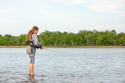 Side view of a young girl standing in the shallow water of a lake as she tries to catch a fish.