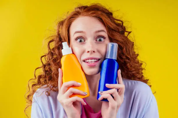 redhaired ginger curly woman holding sunscreen bottle making the right choice in studio yellow background.