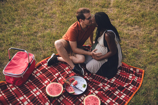 Multiracial couple going for a picnic in a public park, In this photo, husband touching her wife's pregnant belly and giving her a kiss.