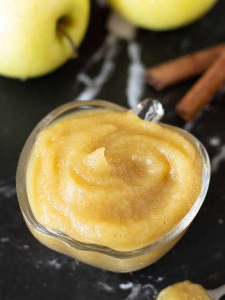 Homemade apple puree in a glass bowl with a swirl on top, cinnamon, and golden delicious apple on a dark table stock photo