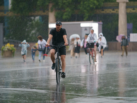 Moscow, Russia - July 10, 2022: Raining day in city at summer. Wet young woman hiding under raincoat and cycling. Man cycling. Texture of strong, fresh, powerful water drops, sprays. Tropical storm.