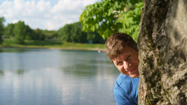 Handsome man resting in the Moscow public park in front of lake. He is hiding behind tree in sunny summer day. He peeking out from behind tree. Concept of the happiness and good mood. Frontal view