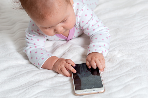 A baby in sliders is lying on the bed and playing with a smartphone. Top view. Concept of children's games with modern devices.