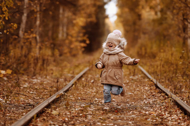 A child girl 6 years old in a beige jacket and a hat walks along the abandoned rails in the autumn forest. She is laughing. Walks in autumn, railway.