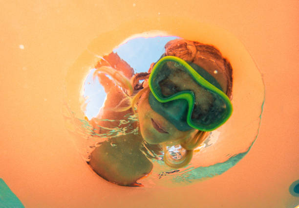 Low angle view of blond hair boy submerging his head though yellow inflatable ring. Playful kid looking at camera.