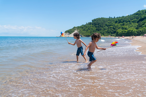 Brothers playing in the waves crashing on the shoreline. They have a colorful weather vane toy in their hands. Shot with a full frame camera on a sunny summer day.