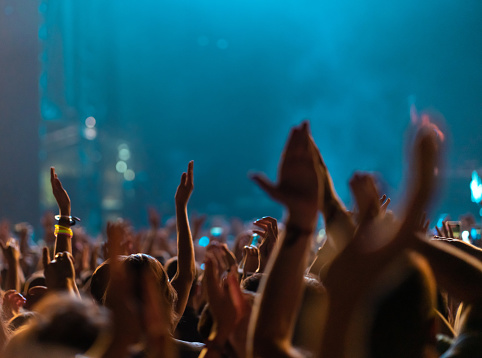 Rear view of a large group of people in front of a music festival stage, under teal stage lights. Crowd is excited and dancing, raising hands, clapping, punching the air, filming with mobile phones, etc...
