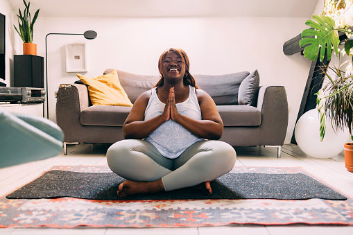 Young Afro-American woman doing yoga at home. She is sitting in a lotus pose, with her eyes closed.