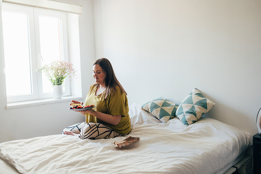 Young Caucasian woman sitting on the bed in her bedroom, eating her breakfast.