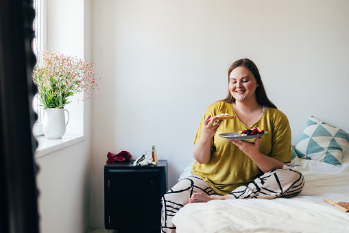 Young Caucasian woman sitting on the bed in her bedroom, eating her breakfast.