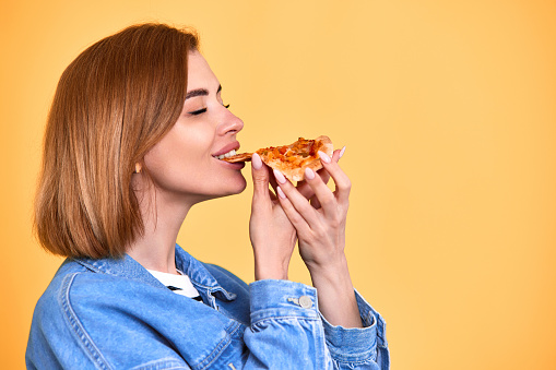 young woman eating with satisfation pizza slice and looking delighted on yellow background. copy space