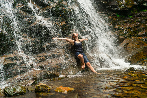 Woman in bikini sit on rock, meditating in yoga pose under falling water of Suwat waterfall in tropical jungle. Nature day tour, adventure at family tourist camp on summer vacation in Bali island