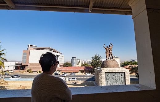 A woman looking at the Genocide Memorial on Robert Mugabe Avenue of Windhoek in Khomas Region, Namibia. She is standing on a free to enter, public area of the Old Fort, which has no charge and is currently disused and locked up. This memorial represents the 'untold hardships and suffering' at the hands of the imperial German Schutztruppe during the 1904–07 war.  It was erected by the North Korean architectural company Mansudae Overseas Projects. Noted as the first genocide of the 20th century, this act against the indigenous Herero and Nama people was overseen by Heinrich Ernst Göring, father of Nazi Luftwaffe chief and Hitler's deputy, Hermann Göring. The statue depicts a man and woman with their arms around each other, symbolising freedom. Ironically Mugabe (after whom the avenue is named) was himself a dictator accused of perpetrating the Gukurahundi genocide in Zimbabwe.