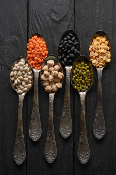 Variety of legumes in old silver spoons on a black wooden background. stock photo