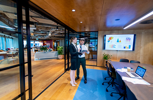 Business Man and woman standing working together on a digital tablet. They are both well dressed standing in a modern office. There is an infographic on a tv showing financial graphs and charts. There is a laptop and paperwork on the table. They are happy and smiling
