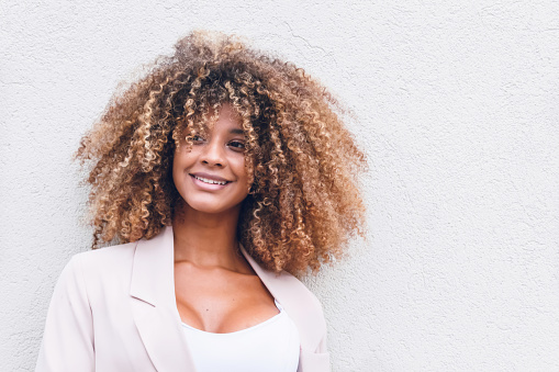 face of beautiful afro woman with curly hair on white wall background in the city smiling happy