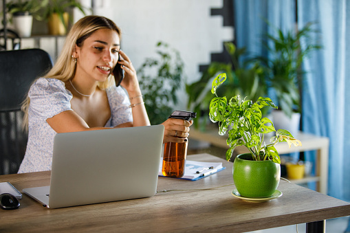 Copy space shot of young female entrepreneur sitting at her desk at the office, talking on the phone and using a spray bottle and spraying a plant with water.