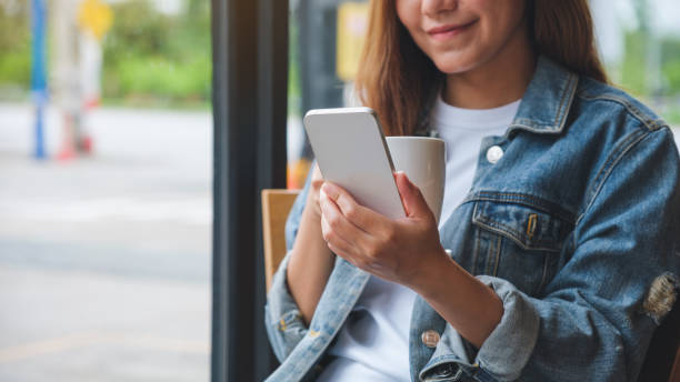 Closeup image of a young woman holding and using mobile phone while drinking coffee in cafe