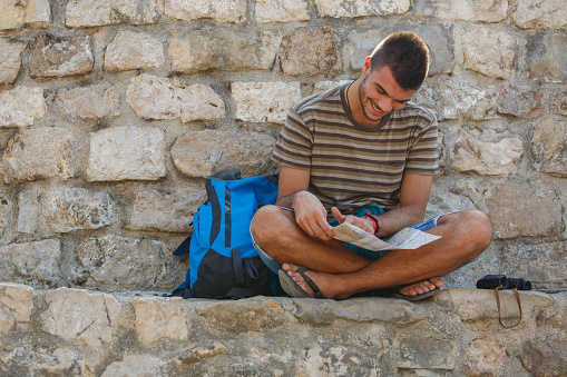 Wide shot of joyful young man sitting on the stone wall, reading a paper map to see where to next on his hike.