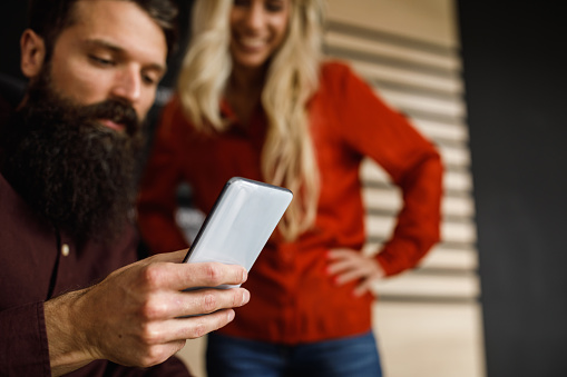 Selective focus shot of hipster businessman showing a funny meme, via smart phone, to a female colleague who is standing by his side with hands on hips. Focus on foreground.