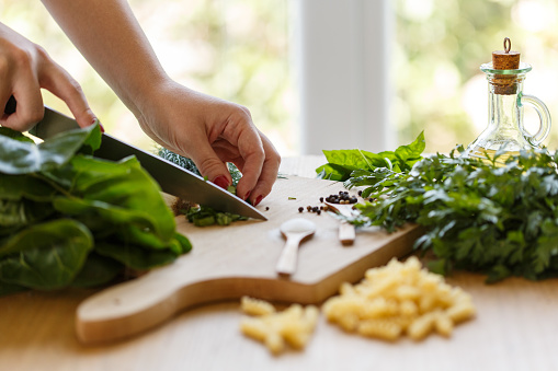 Copy space shot of unrecognizable woman using a kitchen knife and chopping fresh basil on a wooden cutting board when preparing a delicious pasta dish.