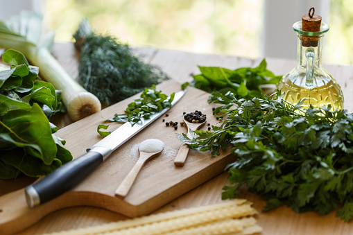 Flat lay of abundance of fresh herbs such as basil, dill, parsley and chard, laid out around a wooden chopping board with salt and pepper in wooden spoons, and kitchen knife.
