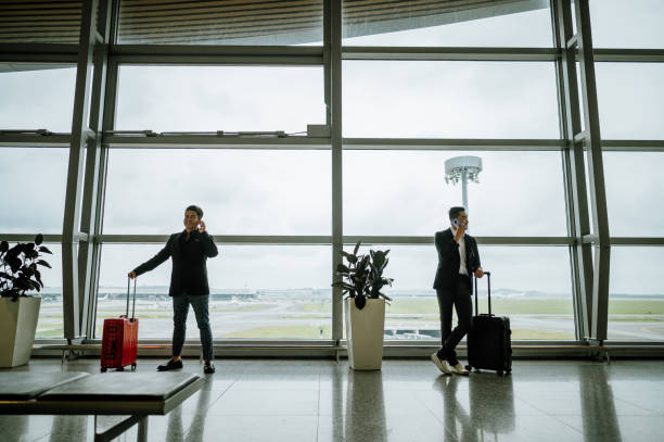 Shot of full length of two Asian Chinese businessmen using smartphone while standing through window of an airport terminal. Full length of two Asian Chinese businessmen using smartphone while standing through window of an airport terminal. kuala lumpur airport stock pictures, royalty-free photos & images