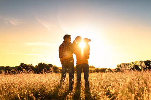 Mother, father, daughter standing in a open field facing nature sunset.