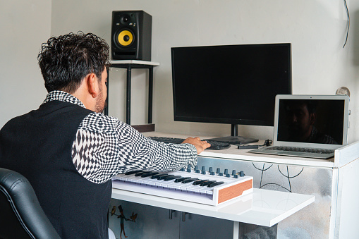 Music producer working in his studio with his computer, laptop, keyboard, and microphone. White room with music producer equipment. Recording and editing music studio. Producer of music dressed with black and white shirt.