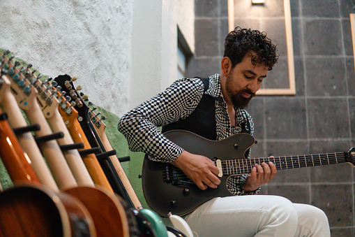 Hispanic musician playing a song with his black guitar sitting next to different guitars on a stand. He is wearing a rocker style with a black and white outfit. He is in a music studio.