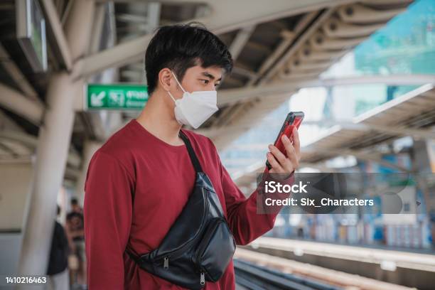 Asian Businessman Passenger Wearing Protective Face Mask Using Mobile Phone And Waiting For Bts Skytrain Or Mrt Underground Train At Railway Platform While Traveling To Work Bangkok Thailand Stock Photo - Download Image Now