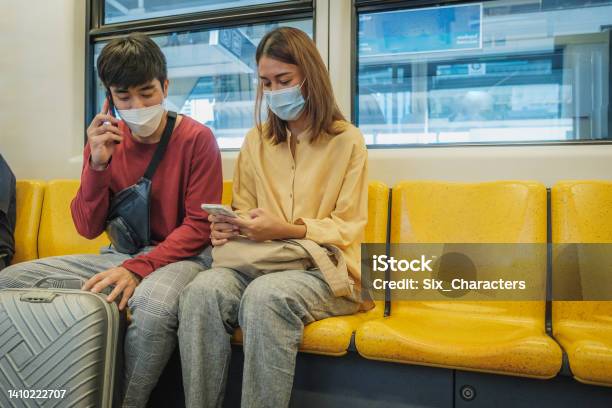 Two Asian Business People Colleague Or Couple Wearing Protective Face Mask Using Mobile Phone And Sitting In Bts Skytrain Or Mrt Underground Train While Traveling To Work Bangkok Thailand Stock Photo - Download Image Now